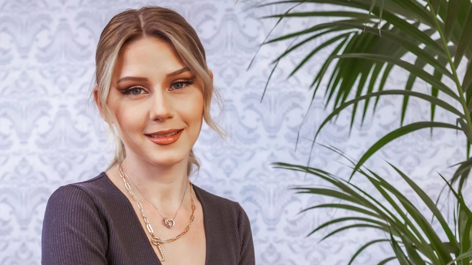 Loredana Wollny lächelt vor einer gemusterten Tapete neben einer Palme